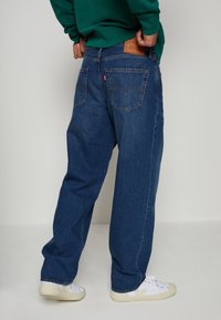 Blå denimjeans med en avslappnad passform, synliga sömmar och två bakfickor. Tan läderlappar på midjan, kombinerat med vita sneakers.