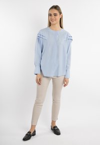 Vaaleansininen paita, jossa on ryppyjä olkapäillä, pitkät hihat ja etupaneeli. Yhdistetty beigen värisiin slim fit -housuihin ja mustiin loafereihin.