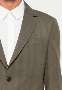 Olivgrön herringbone-blazer med slagkragar, enkel knappstängning och bröstficka. Bärs över en vit skjorta med krage.