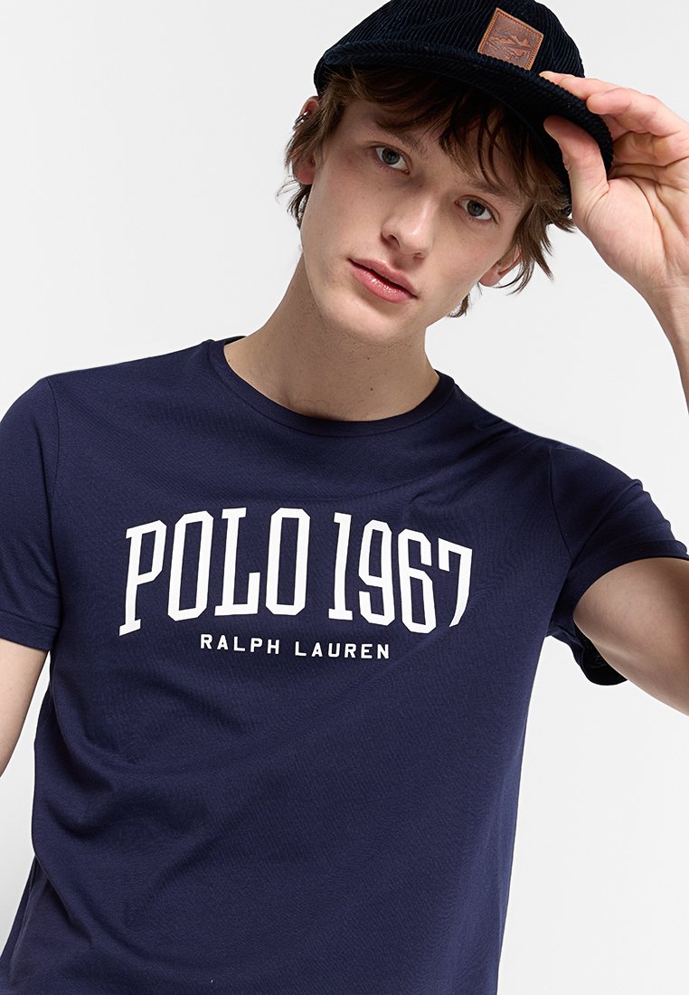 T-shirt in cotone blu navy con grande testo bianco "POLO 1967" e logo "RALPH LAUREN", abbinato a un berretto nero texturizzato con una toppa in pelle.