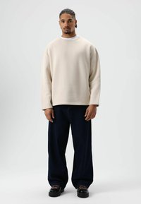 Sweat-shirt beige oversize à col rond, associé à un jean large bleu foncé et des chaussures noires avec des accents roses.