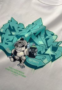 Witte t-shirt met een cartoon gorilla die een spuitbus vasthoudt, omgeven door turquoise graffiti-achtige tekst en sterren, met groene letters.