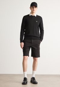 Svart genser med V-hals og logo, hvit skjorte med krage under, svarte cargo-shorts, hvite sokker, svarte sko, og sort caps.