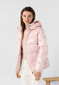 Giacca a vento lucida di color rosa chiaro, con cappuccio, dotata di chiusura con zip sul davanti e polsini elasticizzati. Indossata sopra un top chiaro e abbinata a pantaloni bianchi.