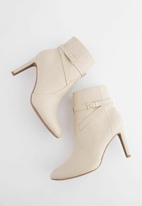 Stivaletti beige alla caviglia con punta appuntita e tacco sottile. Presentano una fascia laterale con fibbia e una texture in pelle liscia.