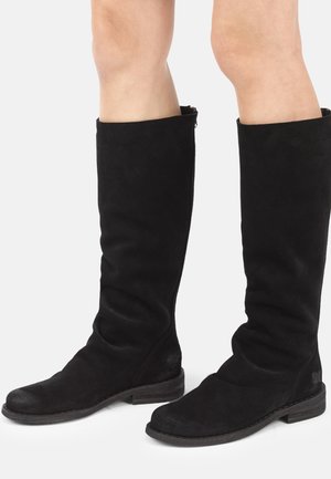 Bottes hautes en daim noir portées sur des jambes nues, avec des talons bas en bois et un design légèrement ample de la tige.