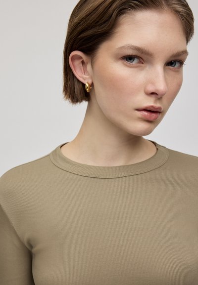 Geribd, langhalstrui in olijfgroen, met een ronde hals. Gouden creolen accentueren een close-up van het gezicht van het model.