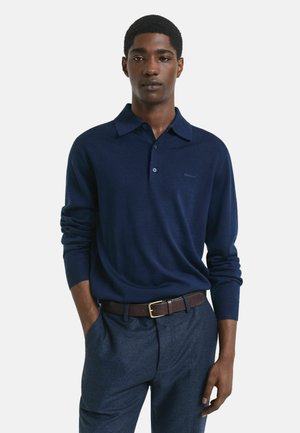 Jongeman in een marineblauwe polo met lange mouwen, ingestopt in een blauwe broek met een leren bruine riem, staand met één hand in zijn zak.
