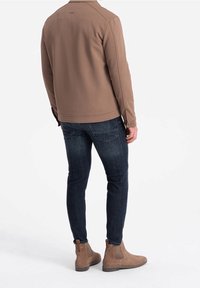 Brun tekstureret jakke, tætsiddende mørke jeans og lysebrune ankelstøvler. Jakken har krave og et subtilt logo på ryggen. Fokus på rene, moderne linjer.