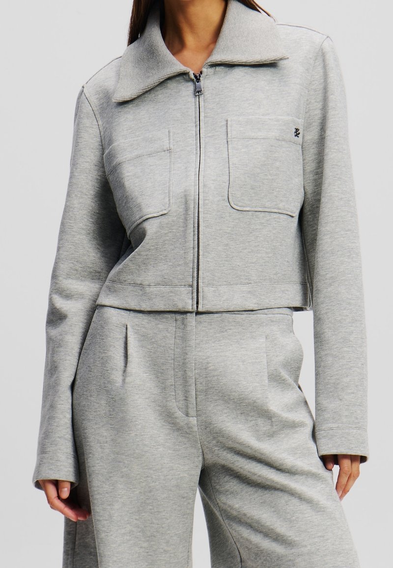 Femme portant une veste courte gris clair zippée avec de grandes poches avant et un pantalon taille haute assorti, sur un fond uni.