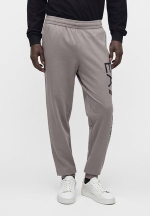 Mand iført grå joggingbukser med sort logo tekst på siden og hvide sneakers, stående mod en ensfarvet lys baggrund.