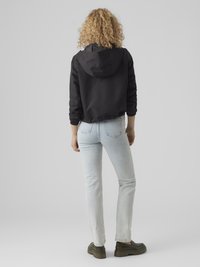 Ženska s kodrastimi lasmi, oblečena v črno jakno s kapuco, svetlo modre kavbojke in zelene robustne čevlje, stoji ob sivem ozadju.