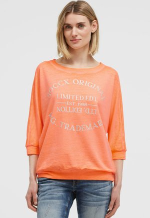 Frau mit kurzen blonden Haaren, die einen lockeren orangenen Pullover mit weißem Text und blaue, abgenutzte Jeans trägt, steht vor einem schlichten Hintergrund.
