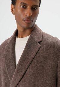 Manteau en laine marron avec un col large, un tissu texturé et une coupe décontractée, porté sur un pull beige clair. Fini lisse et couture notable.