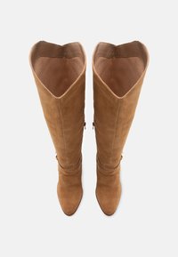 Κόκκινα σουέτ μπότες μέχρι το γόνατο με μυτερή μύτη, διαθέτουν ευρύ άνοιγμα, διακριτικά πτυχές γύρω από τον αστράγαλο και χάλκινα αξεσουάρ.