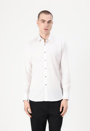 Camisa blanca de manga larga con botones negros, cuello abotonado y una textura suave. Combinada con pantalones negros de diseño minimalista.