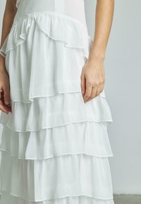 Weißes, geschichtetes Kleid mit Rüschendetails. Aus leichtem Stoff gefertigt, mit einer strukturierten Oberfläche und sichtbaren Nähten entlang der Kanten.