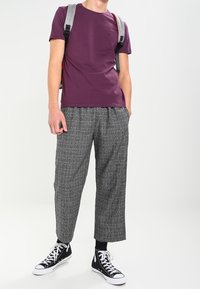 Camiseta de manga corta color marrón, pantalones cortos de cuadros grises, y zapatillas altas negras. Mochila gris llevada sobre un hombro.