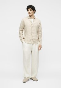 Άνδρας με σγουρά μαύρα μαλλιά, που φοράει μπεζ πουκάμισο από λινό, άνετο άσπρο παντελόνι και μπεζ παπούτσια, στέκεται με τα χέρια στις τσέπες.