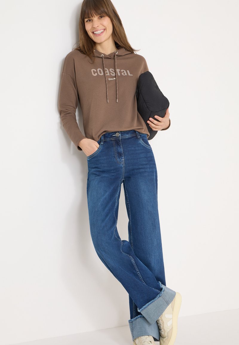 Sweat à capuche marron avec le texte "COASTAL", associé à un jean bleu taille haute aux ourlets effilochés et des baskets beiges. Tenant un objet noir.