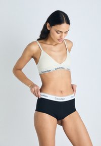 Belo bež bralet Calvin Klein z tankimi naramnicami, v kombinaciji z črnimi visokim pasom kratkimi hlačkami, ki imajo bel značkovni pas.