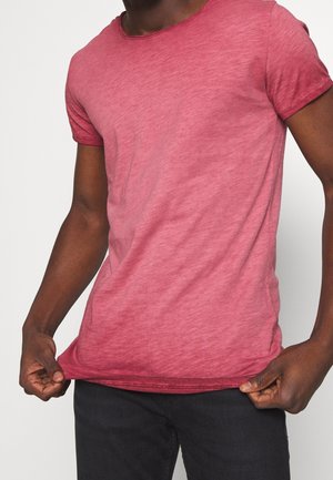 Uomo che indossa una maglietta aderente a maniche corte di colore rosso mélange e pantaloni neri, tenendo l'orlo della maglietta con entrambe le mani.