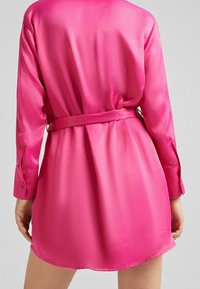 Różowa satynowa sukienka koszulowa z długimi rękawami, z wiązanym pasem i zapinanymi mankietami. Gładka tekstura i delikatny połysk na całej powierzchni.