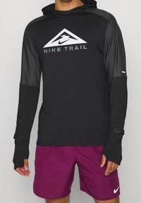 Chemise de sport noire à manches longues avec capuche, comprenant des accents gris, un logo triangulaire et des trous pour les pouces. Associée à un short bordeaux.