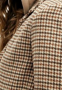 Brun och krämfärgad hundtandsmönstrad tygbit med strukturerad yta, visad på en plaggens axel- och ärmområde.