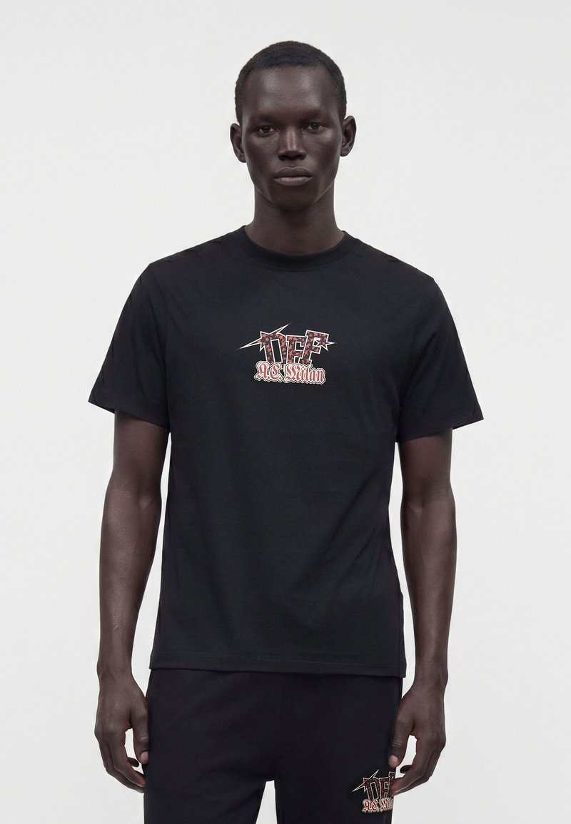 Homme portant un t-shirt noir et un pantalon avec un texte graphique rouge et blanc "DEF AC Milan" à l'avant, debout sur un fond clair uni.