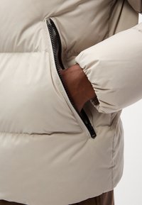 Puffer jacket with a beige exterior, featuring a side pocket with a black zipper and gathered cuffs for a snug fit. Smooth fabric texture.