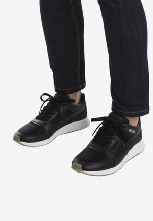 Baskets de sport noires avec une tige texturée, une semelle intermédiaire blanche et des lacets noirs. Présente un logo doré discret sur le côté.
