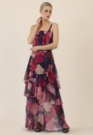 Frau trägt ein mehrlagiges, ärmelloses Maxikleid mit dunklem Blumenmuster in Rot-, Pink- und Lilatönen, steht mit teilweise verschränkten Armen.