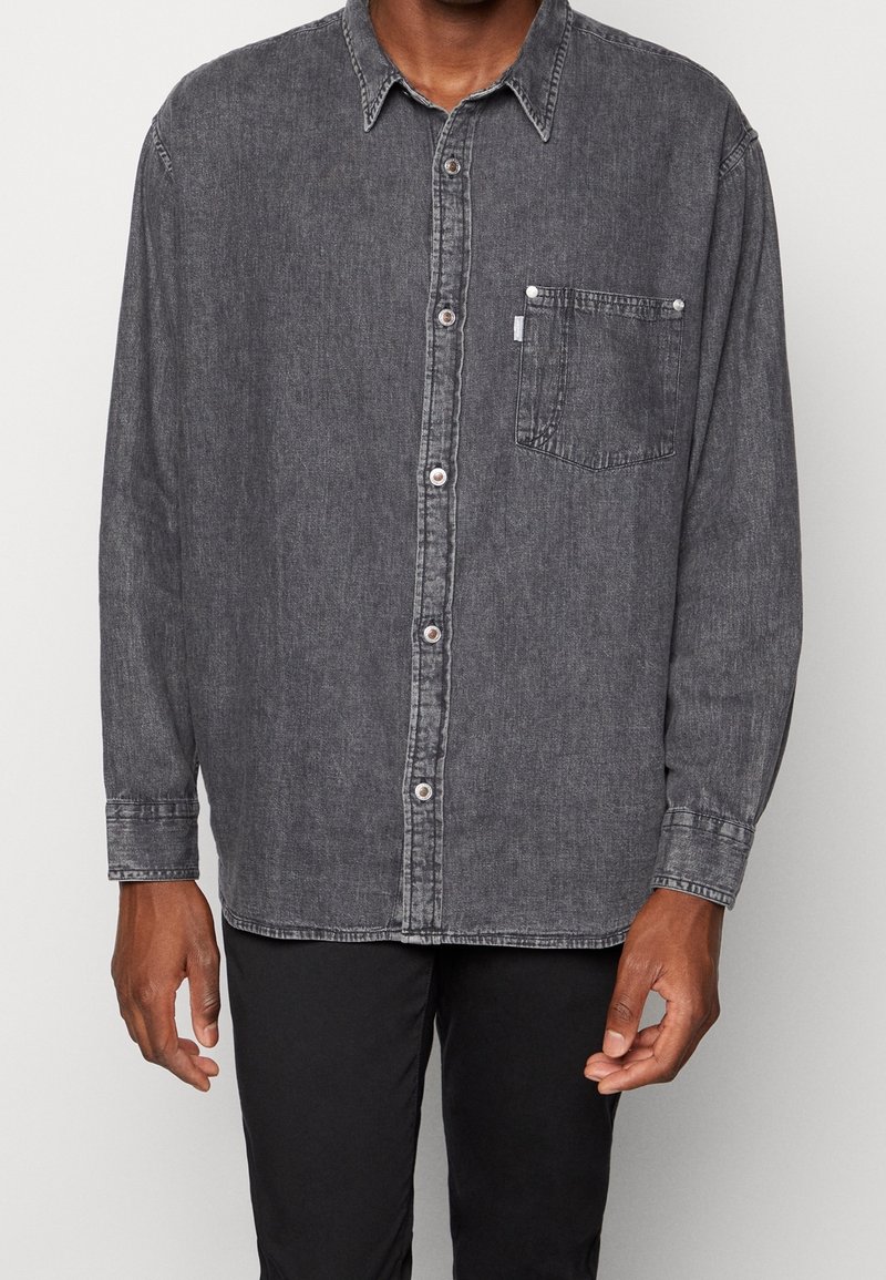 Camicia in denim grigio scuro con bottoni frontali, maniche lunghe e tasca sul petto. Presenta un colletto classico e dettagli di cucitura discreti.