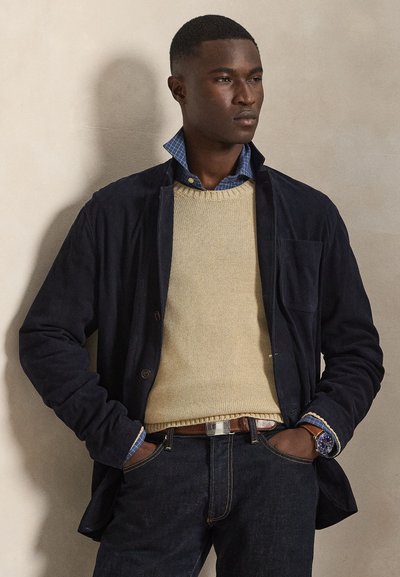 Marineblå semsket skinnjakke over en beige strikkegenser, med en blå rutete skjortekrage, kombinert med mørke denimjeans og en sølvklokke.