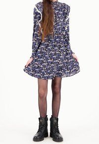 Abito floreale con maniche lunghe, dettagli arricciati e vita aderente. Fiori blu e bianchi su uno sfondo scuro; abbinato a stivali da combattimento neri.