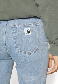 Light blue denim jeans with a slightly tapered fit, featuring a white patch logo on the back pocket and visible stitching details.