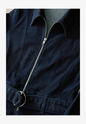 Tumši zili denima kombinezons ar priekšējo rāvējslēdzēju, norādītu apkakli un jostu ar apaļu metāla gredzenu. Gluda tekstūra un piegriezums.