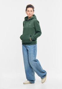 Felpa verde con cappuccio realizzata in tessuto morbido, caratterizzata da una tasca anteriore a marsupio, lacci e polsini a coste, abbinata a jeans blu dalla vestibilità ampia.