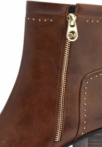 Bottine en simili cuir marron avec fermeture éclair dorée, accents de clous en métal et finition texturée. Elle présente un petit talon carré et un panneau latéral.