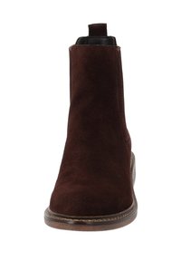 Stivaletto alla caviglia in camoscio marrone con punta tonda, pannelli laterali elastici e un tirante sul retro. Presenta una suola strutturata e cuciture a contrasto.