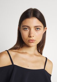 Top preto sem mangas com alças finas, textura suave e um corte largo. O modelo apresenta maquilhagem minimalista e cabelo liso.
