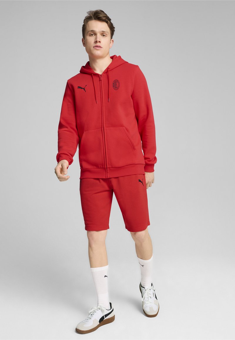 Rød træningsdragt bestående af en hætte-sweatshirt med lynlås og matchende shorts. Har sorte logo-accenter på begge dele. Materialet er en bomuldsblanding.