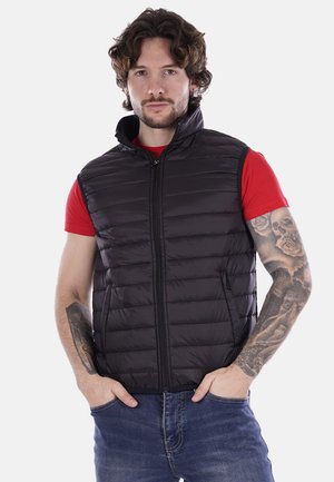 Gilet imbottito nero con collo alto, chiusura frontale con zip e tasche laterali. Indossato sopra una maglietta rossa, con modelli a righe orizzontali.