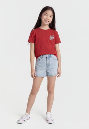 Jongenlijk meisje staand, gekleed in een rood Santa Cruz T-shirt, lichte spijkershorts en witte sneakers, glimlachend tegen een effen witte achtergrond.