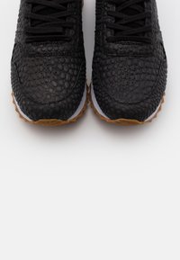 Black textured sneakers with a patterned upper, featuring a rubber sole and traditional lace closure. Accents include a white midsole and gum sole.