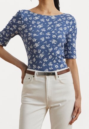 Kobieta ubrana w dopasowaną bluzkę w niebieskie i białe kwiatowe wzory z rękawami do łokcia, wsuniętą w białe spodnie z wysokim stanem i brązowym paskiem.