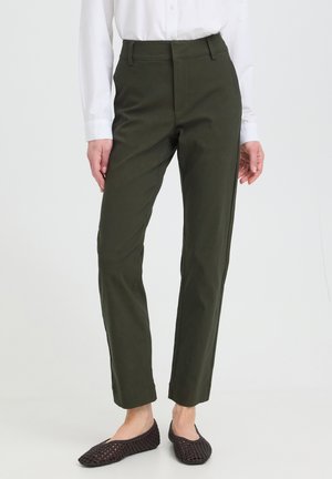 Femme portant un pantalon droit vert foncé, une chemise blanche à manches longues et des chaussures plates tissées foncées, debout devant un fond uni.