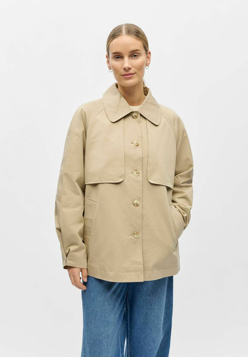 Femme aux cheveux blonds portant une veste beige boutonnée à large col et un jean bleu, debout avec une main dans la poche, sur un fond uni.