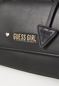 Sac à main noir en faux cuir texturé avec un logo doré "GUESS GIRL", un accent en forme de cœur et une étiquette en forme de triangle.
